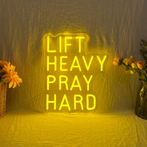 May include: A yellow neon sign with the words "LIFT HEAVY PRAY HARD" in a stacked format. The sign is illuminated against a yellow background, with a woven basket of flowers and a vase of flowers on either side.