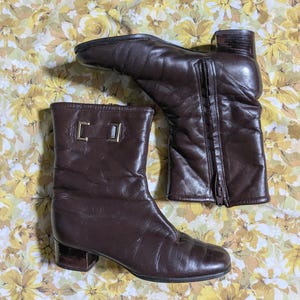 May include: A pair of dark brown leather ankle boots featuring a low block heel, a side zipper closure, and a decorative metallic rectangular detail on the upper shaft. The boots have a smooth finish and a rounded toe design with visible stitching.