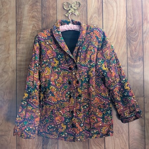 May include: A button-down jacket with a vibrant paisley pattern in shades of orange, purple, green, and yellow. The jacket has a black lining and black buttons. The jacket is hanging on a pink padded hanger.