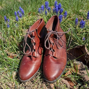 May include: A pair of brown leather ankle boots with laces. The boots are set in a bed of green grass and blue flowers. The boots have a classic design with a rounded toe and a low heel.