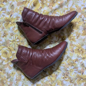 May include: A pair of dark brown leather ankle boots with a soft, ruched texture. The boots feature a pointed toe, a flat sole, and a side zipper closure on the upper boot. The leather has a visible grain and a slightly slouchy, flexible appearance.