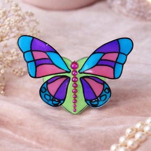 May include: A colorful butterfly-shaped brooch with a heart-shaped base. The wings are divided into sections of blue, pink, and purple. A row of pink beads runs down the center. The brooch is set against a soft pink background.