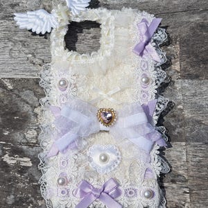 May include: Ivory lace phone case adorned with white wings, a cross, lavender bows, and faux pearls. A heart-shaped jewel is centered on the case. The case has a delicate, feminine aesthetic.