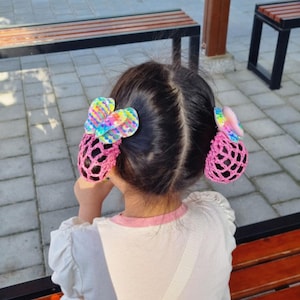 May include: Two hair bun covers featuring a pink crocheted net design and a decorative multi-coloured sequined butterfly accent on each side. The hair accessories are secured over two dark hair buns, providing a textured and colourful appearance.
