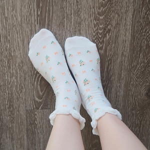 Può includere: Un paio di calzini bianchi con un motivo floreale rosa e verde. I calzini hanno un bordo a balze e sono esposti su una superficie di legno.