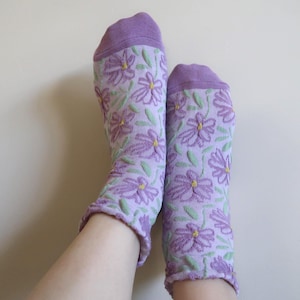 May include: A pair of lavender socks with a floral pattern. The socks feature a repeating design of purple flowers with yellow centers and green leaves. The toes and cuffs are a solid purple color.
