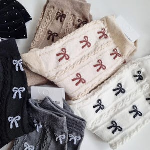 May include: An assortment of socks in various colors, including black, gray, beige, and white. Each pair features a cable knit design and is adorned with embroidered bow accents. Some socks have polka dots. The socks are neatly arranged, showcasing their different styles.