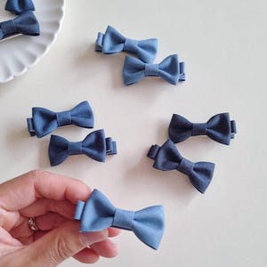 Puede incluir: Varios lazos azules pequeños para el pelo están dispuestos sobre una superficie blanca. Algunos están agrupados en un plato blanco, otros están dispersos. Un lazo es sostenido por una mano. Los lazos tienen un aspecto texturizado.