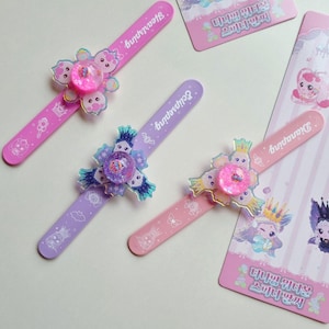 Puede incluir: Tres pulseras de bofetada con motivos de personajes de dibujos animados y elementos giratorios centrales. Una pulsera rosa etiquetada como Heartsping, una morada como Eclipseping y una rosa claro como Dianaping, con tarjetas de presentación a juego.