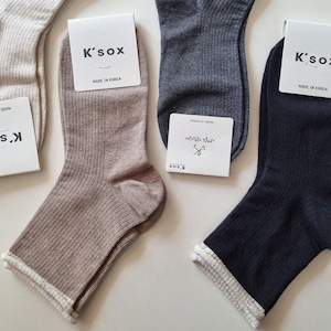 May include: A close-up of several pairs of socks in various colors, including beige, brown, gray, and navy. Each sock has a ribbed texture and a white trim at the cuff. The socks have tags that read "K'sox" and "Made in Korea".
