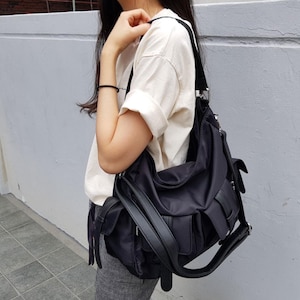 Puede incluir: Bolso de hombro de nailon negro con múltiples bolsillos y una correa ancha ajustable. El bolso tiene forma curva y cierre de solapa. El bolso se lleva al hombro.