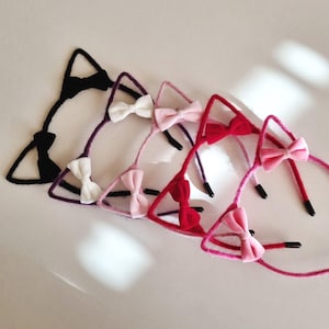 May include: A selection of cat ear headbands in a range of colours. Each headband has a triangular ear design and a small bow. The colours include black, purple, white, pink, and red. The headbands are arranged in a line.
