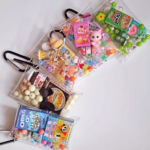 May include: Several clear plastic pouches filled with miniature items, including snacks, candies, and small toys. Each pouch has a black carabiner clip attached. The contents include Oreo O's cereal boxes, a Nutella jar, and a pink cat figurine.