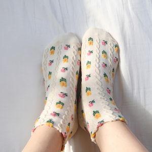 May include: A pair of cream-colored ankle socks featuring a repeating pattern of small yellow and pink flowers with green stems. The socks have a vertical cable knit texture running along the sides and a ribbed cuff at the top edge.