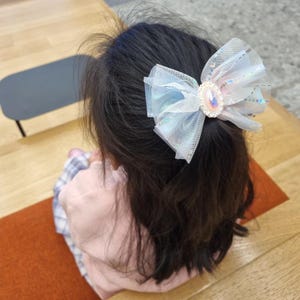 Könnte beinhalten: Eine hellblaue und weiße Haarschleife mit einem zentralen irisierenden Edelstein ist an dunklem Haar befestigt. Die Schleife besteht aus durchsichtigem, glitzerndem Stoff und ist ein zartes und skurriles Accessoire.