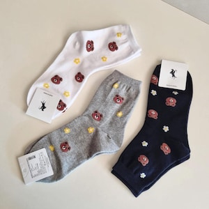 Puede incluir: Tres pares de calcetines en blanco, gris y azul marino, cada uno adornado con un patrón repetido de caras de oso marrón y flores amarillas o blancas. Cada calcetín tiene una etiqueta adjunta.