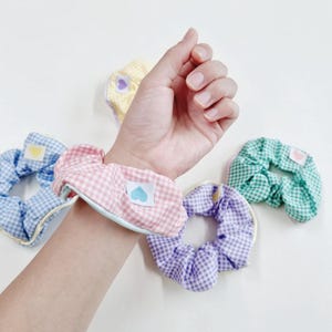 May include: A collection of colorful gingham scrunchies. One pink scrunchie is worn on a wrist. Each scrunchie has a small heart-shaped patch. Colors include pink, blue, green, yellow, and purple. The scrunchies are made of fabric.