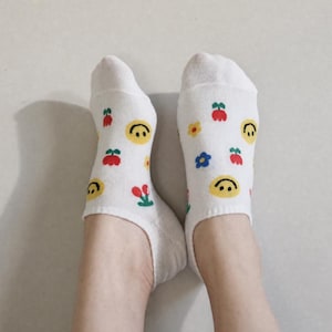May include: A pair of white ankle socks featuring a repeating pattern of yellow smiley faces, red cherries, and small blue and yellow flowers. The socks have a low-cut design that sits below the ankle, with a consistent knit texture throughout the fabric.