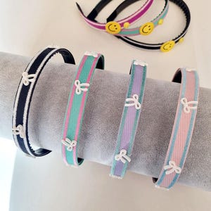 May include: A collection of headbands in various colors, including black and white, teal and pink, lavender and white, and pink and blue. Each headband features a repeating bow design. Some headbands have smiley face accents.