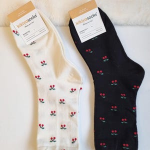 Puede incluir: Dos pares de calcetines de mujer, uno marfil y otro negro, cada uno adornado con un patrón repetido de pequeñas cerezas rojas con hojas verdes. Los calcetines tienen etiquetas que dicen "kikiyasocks". El par marfil está etiquetado como "Ivory", el negro como "Black".