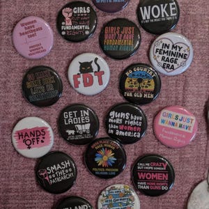 May include: A selection of circular badges with feminist messages and designs. The badges display slogans such as "Girls Just Want to Have Fundamental Rights," "Smash the Patriarchy," and "Women have heartbeats too!"
