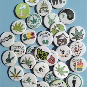 Op de afbeelding: Een verzameling ronde, witte buttons met diverse cannabis-thema's. De buttons tonen afbeeldingen van marihuanabladeren, rookattributen en tekst zoals "4:20", "Roll Up" en "Get Lit". De achtergrond is een lichtblauwe ondergrond.