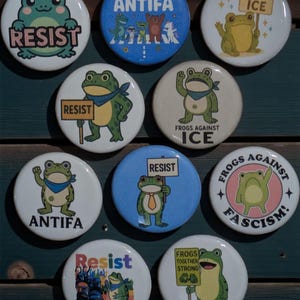 May include: A collection of round buttons with frog illustrations and various text. The buttons feature phrases like "RESIST", "ANTIFA", "ABOLISH ICE", and "FROGS AGAINST FASCISM". The frogs are depicted in different poses and styles.