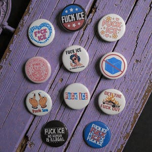 Puede incluir: Una colección de botones redondos, blancos y negros con varios diseños gráficos y texto. Los botones presentan frases como "FUCK ICE" y "NO HUMAN IS ILLEGAL". Los botones están dispuestos sobre una superficie de madera morada desgastada.