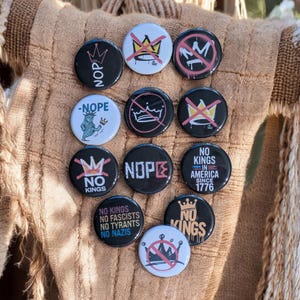 Può includere: Una collezione di bottoni bianchi e neri con vari slogan e grafiche anti-monarchia. I bottoni presentano frasi come "NOPE", "NO KINGS" e "NO KINGS IN AMERICA SINCE 1776". Alcuni bottoni hanno disegni di corone sbarrate.