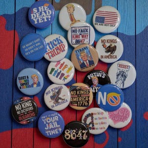 Puede incluir: Una colección de botones redondos con varios mensajes y gráficos políticos. Los botones presentan textos como "No Faux King Way", "No Kings in America" y "Resist". La paleta de colores incluye rojo, blanco, azul y negro.