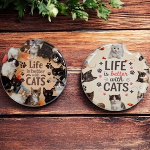 May include: Two round car coasters with cat designs. One coaster has a black background with various cat images and the text "Life is better with cats." The other coaster has a cream background with cat images and the same text.