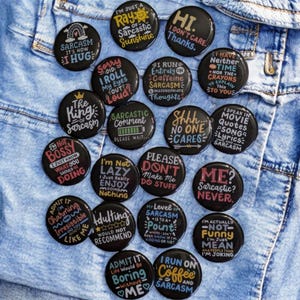 May include: A collection of black, round buttons with white and red text, displaying humorous sarcastic phrases. The buttons are arranged on a denim background. Phrases include "Sarcasm It's How I Hug" and "I'm Not Bossy I Just Know What You Should Be Doing."