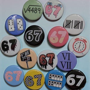 May include: A collection of round pinback buttons with various designs. Some buttons feature the number "67" in different styles, while others display images like a dominoes, a square root symbol, and an alarm clock. The color palette includes pink, blue, black, and yellow.