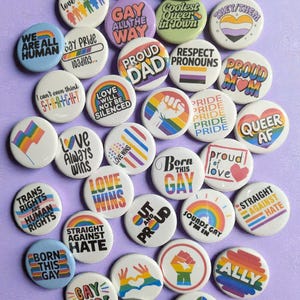 May include: A collection of round, colorful pride buttons with various slogans and designs. The buttons feature phrases like "Love is Love," "Gay Pride," "Proud Dad," and "Queer AF." Rainbow and gender symbols are also present.