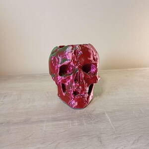 May include: A red skull-shaped decorative object with a metallic finish. The skull has visible eye sockets, a nose cavity, and a detailed jawline. The top of the skull is open, suggesting it could be used as a planter or container. The surface reflects green and pink hues.