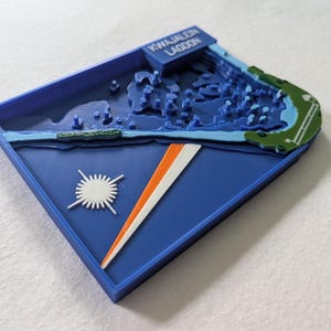 May include: A 3D printed topographic map of Kwajalein Lagoon in blue, green, white, and orange. The piece features a raised relief of the lagoon, a white starburst symbol, and an orange and white diagonal stripe on a dark blue base.