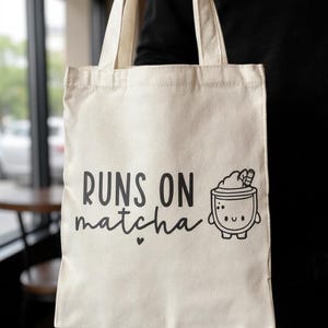 May include: Cream-colored tote bag with black text that reads "RUNS ON matcha" and a cute matcha drink illustration. The bag has two handles and is held up by a person.