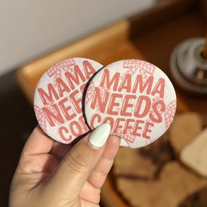 Könnte beinhalten: Zwei runde Untersetzer mit dem Text "MAMA NEEDS COFFEE" in roten Buchstaben. Die Untersetzer haben einen weißen Hintergrund mit einem rosa Vichy-Muster. Die Untersetzer werden in einer Hand gehalten.