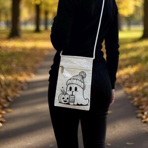 Op de afbeelding: Een crèmekleurige crossbodytas met een zwarte afbeelding van een spook met een gebreide muts, een koffiekopje en een pompoen. De tas heeft een ritssluiting en een witte band. De achtergrond is een vage buitenscène.