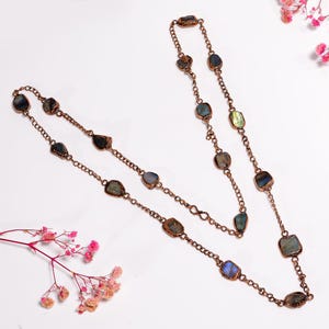 May include: A long necklace featuring a copper-toned metal chain interspersed with multiple irregular, raw-cut labradorite stones. The stones display dark grey and blue hues with iridescent flashes, secured within copper settings along the chain links.