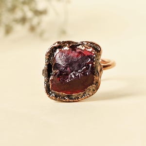Op de afbeelding: Een ruwe granaatring met een koperen band. De granaat heeft een dieprode kleur met een ruw, getextureerd oppervlak. De koperen band heeft een warme, roodbruine tint. De ring is een uniek sieraad.