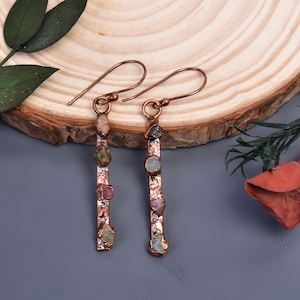 May include: A pair of copper-toned earrings with a vertical bar design. Each earring features a series of colourful, irregularly shaped gemstones in shades of green, pink, and blue. The earrings have a simple hook closure.