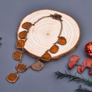 Può includere: Una collana color rame con pietre ambrate di forma irregolare. Le pietre sono incastonate in un metallo color rame e collegate da una delicata catena. La collana è esposta su una fetta di legno su sfondo grigio.