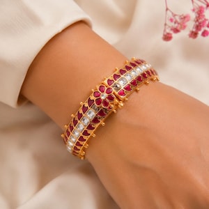Pode incluir: Uma pulseira rígida articulada em tom dourado com uma fila central de pedras quadradas transparentes, ladeada por duas filas de pedras vermelhas em forma de coração. A pulseira inclui um fecho floral com uma pedra central clara.