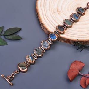Puede incluir: Una pulsera de tono cobrizo con piedras ovaladas de labradorita. Las piedras muestran tonos iridiscentes azules, verdes y grises. La pulsera tiene un cierre de palanca y se exhibe sobre una superficie gris con elementos de madera y naturales.