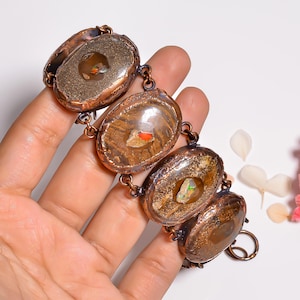 May include: A link bracelet featuring four oval-shaped copper settings, each containing a polished brown stone with a central iridescent opal inlay. The copper frames have a textured, dark patina finish and are connected by small circular copper rings.