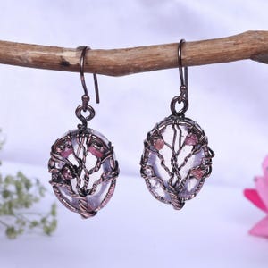May include: A pair of handcrafted earrings featuring oval-shaped, clear gemstones with a tree-of-life design crafted from dark metal wire. Small pink gemstones are embedded within the wire tree. The earrings hang from a dark metal hook.