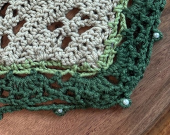 May include: A handmade crochet shawl in shades of green and tan, adorned with small blue flower accents and white bead embellishments. The shawl has a triangular shape and a delicate, openwork design, perfect for a cozy accessory.