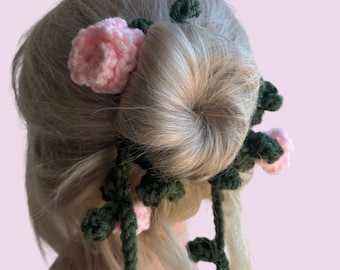 May include: A handmade hair accessory featuring a bun adorned with crocheted pink flowers and green leaves. The floral design is attached to the hair with a dark green vine-like structure, creating a whimsical and natural look.