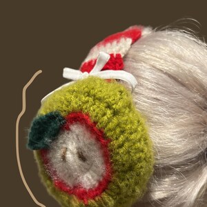 May include: A handmade, crocheted apple-shaped hair accessory. The apple is primarily green with a red outline, white center, and a small green leaf. A red and white striped band and white ribbon complete the design.
